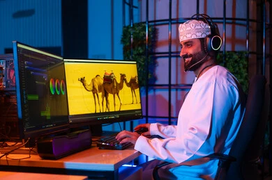 مفهوم صناعة المحتوى الرقمي،تعديل المشاهد وإضافة المؤثرات، صورة من الجانب لمونتير عربي خليجي عماني يرتدي الدشداشة و الكمة وسماعات الرأس يجلس داخل الاستوديو يستخدم شاشات متعددة لتصميم الفيديوهات،  النقر على لوحه المفاتيح