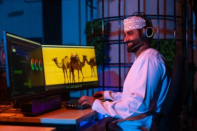 تعديل المشاهد وإضافة المؤثرات، صورة من الجانب لمونتير عربي خليجي عماني يرتدي الدشداشة و الكمة وسماعات الرأس يجلس داخل الاستوديو يستخدم شاشات متعددة لتصميم الفيديوهات، مفهوم صناعة المحتوى الرقمي، النقر على لوحه المفاتيح