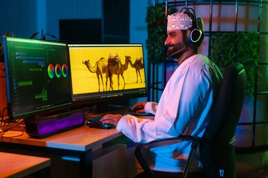 تعابير السعادة والسرور، تعديل المشاهد وإضافة المؤثرات، صورة من الجانب لمونتير عربي خليجي عماني يرتدي الدشداشة و الكمة وسماعات الرأس يجلس داخل الاستوديو يستخدم شاشات متعددة لتصميم الفيديوهات، مفهوم صناعة المحتوى الرقمي، النقر على لوحه المفاتيح