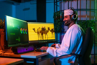 استخدام التقنيات الحديثة ، تعديل المشاهد وإضافة المؤثرات، صورة من الجانب لمونتير عربي خليجي عماني يرتدي الدشداشة و الكمة وسماعات الرأس يجلس داخل الاستوديو يستخدم شاشات متعددة لتصميم الفيديوهات، مفهوم صناعة المحتوى الرقمي، النقر على لوحه المفاتيح