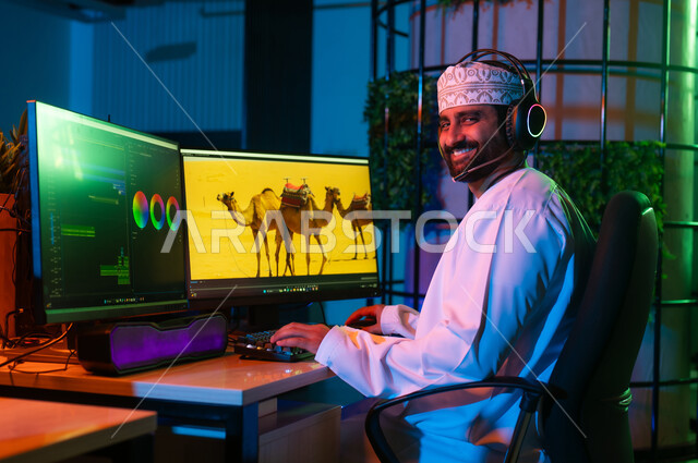 صورة من الجانب لمونتير عربي خليجي عماني يرتدي الدشداشة و الكمة وسماعات الرأس يجلس داخل الاستوديو يستخدم شاشات متعددة لتصميم الفيديوهات، تعديل المشاهد وإضافة المؤثرات، مفهوم صناعة المحتوى الرقمي، النقر على لوحه المفاتيح