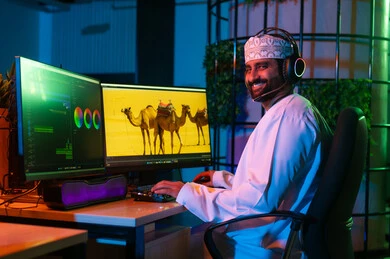 صورة من الجانب لمونتير عربي خليجي عماني يرتدي الدشداشة و الكمة وسماعات الرأس يجلس داخل الاستوديو يستخدم شاشات متعددة لتصميم الفيديوهات، تعديل المشاهد وإضافة المؤثرات، مفهوم صناعة المحتوى الرقمي، النقر على لوحه المفاتيح