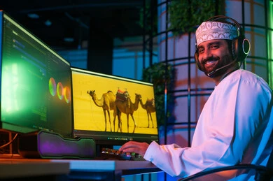 النظر الي الكاميرا بتعابير السعادة ، تعديل المشاهد وإضافة المؤثرات، صورة من الجانب لمونتير عربي خليجي عماني يرتدي الدشداشة و الكمة وسماعات الرأس يجلس داخل الاستوديو يستخدم شاشات متعددة لتصميم الفيديوهات، مفهوم صناعة المحتوى الرقمي، النقر على لوحه المفاتيح