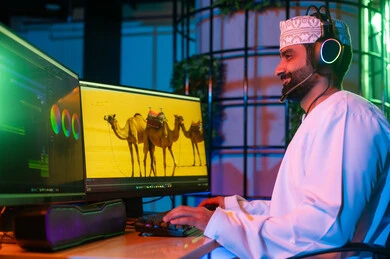 النقر على لوحه المفاتيح، تعديل المشاهد وإضافة المؤثرات، صورة من الجانب لمونتير عربي خليجي عماني يرتدي الدشداشة و الكمة وسماعات الرأس يجلس داخل الاستوديو يستخدم شاشات متعددة لتصميم الفيديوهات، مفهوم صناعة المحتوى الرقمي