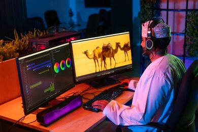 مفهوم صناعة المحتوى الرقمي، تعديل المشاهد وإضافة المؤثرات، صورة من الجانب لمونتير عربي خليجي عماني يرتدي الدشداشة و الكمة وسماعات الرأس يجلس داخل الاستوديو يستخدم شاشات متعددة لتصميم الفيديوهات، النقر على لوحه المفاتيح