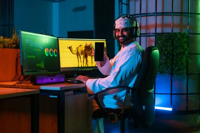 استخدام الهاتف المحمول  ، مفهوم صناعة المحتوى الرقمي، صورة من الجانب لمونتير عربي خليجي عماني يرتدي الدشداشة و الكمة وسماعات الرأس يجلس داخل الاستديو يعمل على ترتيب المشاهد باستخدام برامج الحاسوب والجوال المتطورة  ، ايماءات التفكير و التركيز