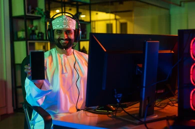 Using a mobile phone, the concept of visual content creation, creativity in the world of editing, a side image of an Arab Gulf Omani editor wearing a dishdasha and kumma with headphones sitting in front of a laptop screen producing a cinematic scene, gestures of immersion and focus.