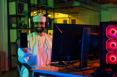 عرض شاشة سوداء فارغة ، مفهوم صناعة المحتوى المرئي، الإبداع في عالم المونتاج ، صورة من الجانب لمونتير عربي خليجي عماني يرتدي الدشداشة والكمة و سماعات الراس يجلس أمام شاشة حاسب محمول يقوم باخراج مشهد سينمائي ، ايماءات الاندماج و التركيز