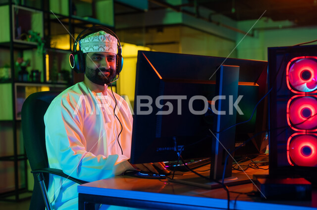 مفهوم صناعة المحتوى المرئي، الإبداع في عالم المونتاج ، صورة من الجانب لمونتير عربي خليجي عماني يرتدي الدشداشة والكمة  و سماعات الراس يجلس أمام شاشة حاسب محمول يقوم باخراج مشهد سينمائي ، ايماءات الاندماج و التركيز