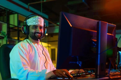 مفهوم الإبداع في عالم المونتاج وصناعة المحتوى المرئي، صورة من الجانب لمونتير عربي خليجي عماني يرتدي الدشداشة والكمة  و سماعات الراس يجلس أمام شاشة حاسب محمول يقوم باخراج مشهد سينمائي ، ايماءات الاندماج و التركيز