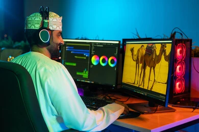 صورة من الجانب لمونتير عربي خليجي عماني يرتدي الدشداشة و الكمة وسماعات الرأس يجلس داخل الاستوديو يستخدم شاشات متعددة لتصميم الفيديوهات، مفهوم صناعة المحتوى الرقمي، تعديل المشاهد وإضافة المؤثرات، النقر على لوحه المفاتيح