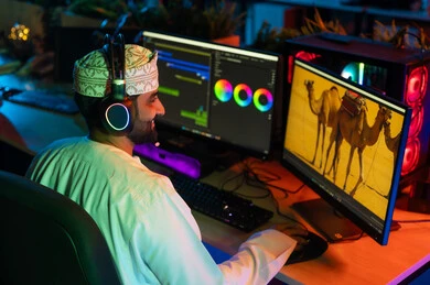 النقر على لوحه المفاتيح، تعديل المشاهد وإضافة المؤثرات، صورة من الجانب لمونتير عربي خليجي عماني يرتدي الدشداشة و الكمة وسماعات الرأس يجلس داخل الاستوديو يستخدم شاشات متعددة لتصميم الفيديوهات، مفهوم صناعة المحتوى الرقمي