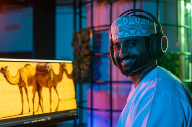صورة من الجانب لمونتير عربي خليجي عماني يرتدي الدشداشة و الكمة وسماعات الرأس يجلس داخل الاستوديو يستخدم شاشات متعددة لتصميم الفيديوهات، تعديل المشاهد وإضافة المؤثرات، مفهوم صناعة المحتوى الرقمي، النقر على لوحه المفاتيح