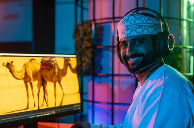 Looking at the camera with expressions of happiness, editing scenes and adding effects, a side image of an Arab Gulf Omani editor wearing a dishdasha and kumma with headphones sitting inside the studio using multiple screens to design videos, the concept of digital content creation, clicking on the keyboard.