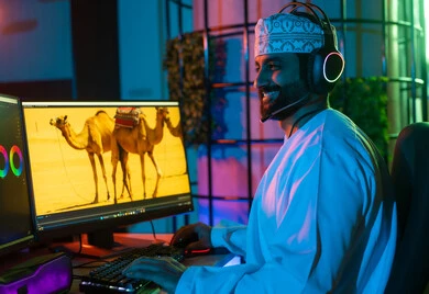 النقر على لوحه المفاتيح، تعديل المشاهد وإضافة المؤثرات، صورة من الجانب لمونتير عربي خليجي عماني يرتدي الدشداشة و الكمة وسماعات الرأس يجلس داخل الاستوديو يستخدم شاشات متعددة لتصميم الفيديوهات، مفهوم صناعة المحتوى الرقمي