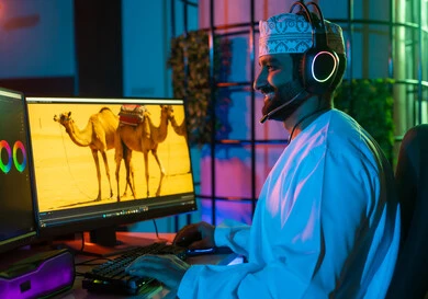 The concept of digital content creation, editing scenes and adding effects, a side image of an Arab Gulf Omani editor wearing a dishdasha and kumma with headphones sitting inside the studio using multiple screens to design videos, clicking on the keyboard.