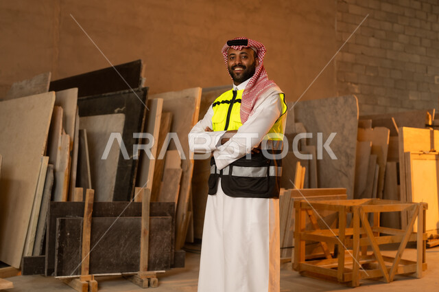 الوقوف بإيدي متقاطعة بتعابير السرور، ورشات عمل في المملكة العربية السعودية, أدوات ومعدات مصنوعة من الرخام، جودة الانتاج المحلي الوطني, صناعة الأثاث المنزلي والمكتبي، رجل مهندس عربي خليجي سعودي يرتدي الثوب التقليدي وزي العمل ينظر الى شيء ما بايماءات السعادة