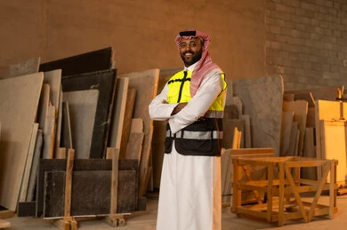 Standing with crossed hands with happy expressions, workshops in Saudi Arabia, tools and equipment made of marble, quality of local national production, home and office furniture industry, a Saudi Arabian Gulf Arab engineer man wearing traditional thobe and work clothes looking at something with happy gestures