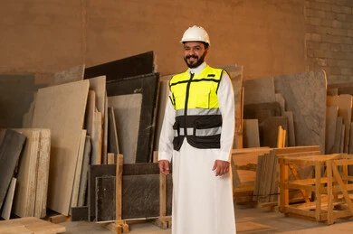 جودة الانتاج المحلي الوطني, صناعة الأثاث المنزلي والمكتبي، رجل مهندس عربي خليجي سعودي يرتدي الثوب التقليدي وزي العمل ينظر الى الكاميرا بايماءات السعادة، الوقوف باستقامة بتعابير السرور، ورشات عمل في المملكة العربية السعودية, أدوات ومعدات مصنوعة من الرخام