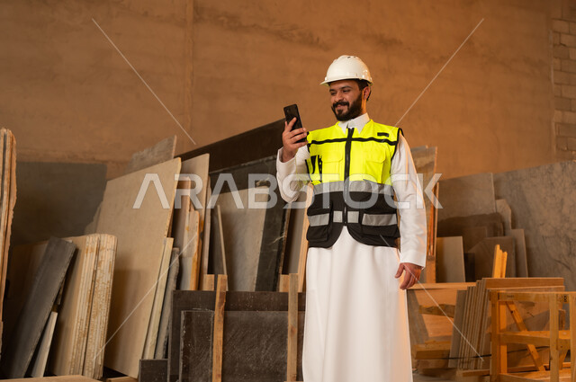 العمل على الهاتف المحمول، ورشات عمل في المملكة العربية السعودية, أدوات ومعدات مصنوعة من الرخام، جودة الانتاج المحلي الوطني, صناعة الأثاث المنزلي والمكتبي، رجل مهندس عربي خليجي سعودي يرتدي الثوب التقليدي وزي العمل يمسك بيده الجوال