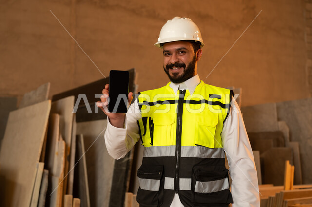 Blank black screen on mobile phone, workshops in Saudi Arabia, tools and equipment made of marble, quality of national local production, home and office furniture industry, a Saudi Arabian Gulf Arab engineer man wearing traditional thobe and work clothes holding a mobile phone in his hand