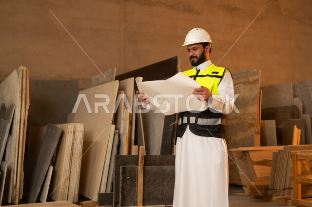 العمل على المخططات والاوراق الهندسية، رجل مهندس عربي خليجي سعودي يرتدي الثوب التقليدي وزي العمل يمسك بيده الورقة، ورشات عمل في السعودية، أدوات ومعدات مصنوعة من الرخام، جودة الانتاج المحلي الوطني, صناعة الأثاث المنزلي والمكتبي