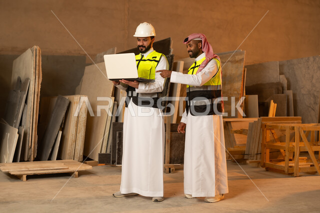 رجلان مهندسان عربيان خليجيان سعوديان يرتديان الثوب التقليدي وزي العمل يعملان على الحاسب المحمول، دمج التقنية والتكنولوجيا في المعامل، ورشات عمل في السعودية، أدوات ومعدات مصنوعة من الرخام، جودة الانتاج المحلي الوطني, صناعة الأثاث المنزلي والمكتبي