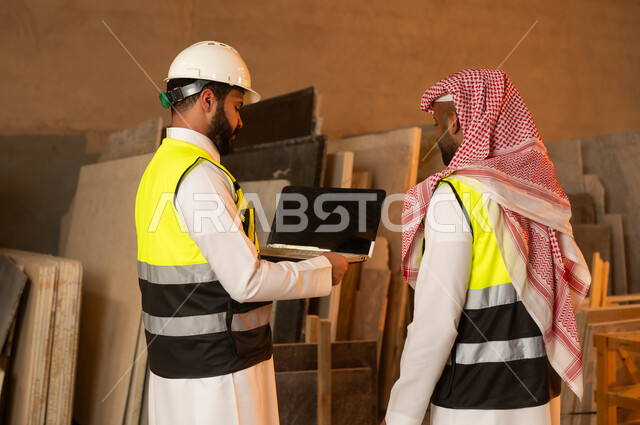 أدوات ومعدات مصنوعة من الرخام، جودة الانتاج المحلي الوطني, صناعة الأثاث المنزلي والمكتبي، صورة من الخلف لرجلين مهندسين عربيين خليجيين سعوديين يرتديان الثوب التقليدي وزي العمل يعملان على الحاسب المحمول، دمج التقنية والتكنولوجيا في المعامل، ورشات عمل في السعودية