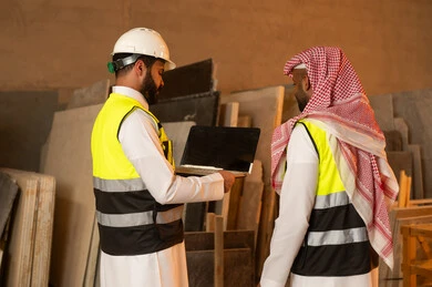 Marble tools and equipment, quality local production, home and office furniture manufacturing, back view of two Saudi Arabian Gulf engineers wearing traditional thobes and work clothes working on laptops, integration of technology in laboratories, workshops in Saudi Arabia