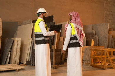 أدوات ومعدات مصنوعة من الرخام، جودة الانتاج المحلي الوطني, صناعة الأثاث المنزلي والمكتبي، صورة من الخلف لرجلين مهندسين عربيين خليجيين سعوديين يرتديان الثوب التقليدي وزي العمل يعملان على الحاسب المحمول، دمج التقنية والتكنولوجيا في المعامل، ورشات عمل في السعودية