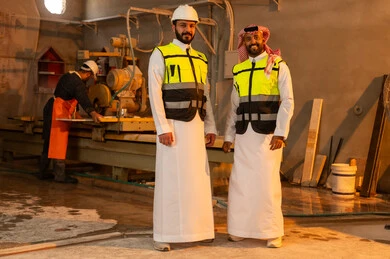 Standing up straight with a happy expression, workshops in Saudi Arabia, tools and equipment made of marble, quality of local national production, home and office furniture industry, a Saudi Arabian Gulf Arab engineer man wearing traditional thobe and work clothes looking at the camera with his friend