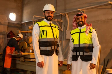 Standing up straight with a happy expression, workshops in Saudi Arabia, tools and equipment made of marble, quality of local national production, home and office furniture industry, a Saudi Arabian Gulf Arab engineer man wearing traditional thobe and work clothes looking at the camera with his friend