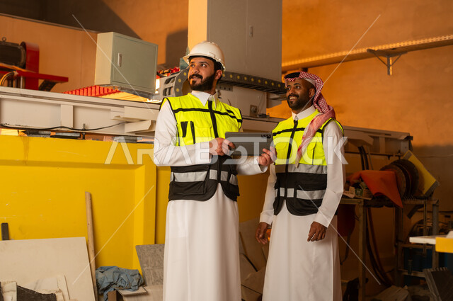 ورشات عمل في المملكة العربية السعودية, أدوات ومعدات مصنوعة من الرخام، جودة الانتاج المحلي الوطني, صناعة الأثاث المنزلي والمكتبي، رجلان مهندسان عربيان خليجيان سعوديان يرتديان الثوب التقليدي وزي العمل يعملان على جهاز التابلت، دمج التقنية والتكنولوجيا في المعامل