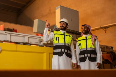 Pointing to something, workshops in Saudi Arabia, tools and equipment made of marble, quality of local national production, old traditional professions and crafts in the Kingdom, home and office furniture manufacturing, a Saudi Arabian Gulf Arab engineer wearing traditional clothing and work clothes standing inside the factory with his colleague