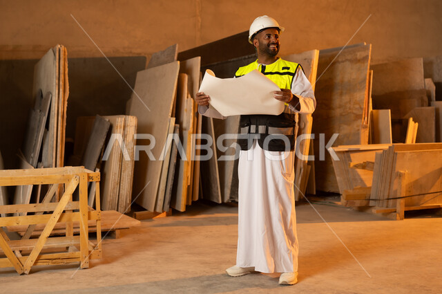 Working on engineering plans and papers, an Arab Gulf Omani engineer wearing a dishdasha and work clothes holding a paper in his hand, workshops in the Sultanate of Oman, tools and equipment made of marble, quality of local national production, home and office furniture manufacturing