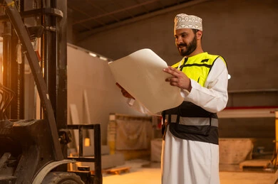 Making a local national product, making home and office furniture, working on engineering plans and papers, an Arab Gulf Omani engineer wearing a dishdasha and work clothes holding a paper in his hand, workshops in the Sultanate of Oman, tools and equipment made of marble