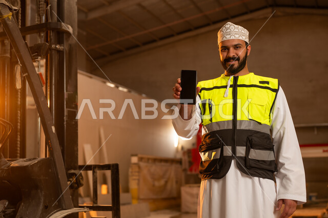 Showing a blank black screen on a mobile phone, workshops in the Sultanate of Oman, tools and equipment made of marble, quality of local national production, home and office furniture manufacturing, an Arab Gulf Omani engineer man wearing a dishdasha and work clothes holding a mobile phone in his hand