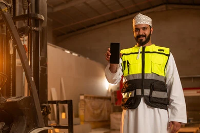 Showing a blank black screen on a mobile phone, workshops in the Sultanate of Oman, tools and equipment made of marble, quality of local national production, home and office furniture manufacturing, an Arab Gulf Omani engineer man wearing a dishdasha and work clothes holding a mobile phone in his hand