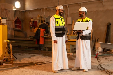 Home and office furniture manufacturing, two Omani Gulf Arab engineers wearing dishdashas and work clothes working on laptops, integrating technology into laboratories, workshops in the Sultanate of Oman, tools and equipment made of marble, quality of local national production