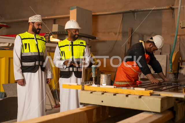 Monitoring and evaluating worker performance via tablet, factories and workshops in the Sultanate of Oman, marble manufacturing machines and equipment, quality of local national production, two Omani Gulf Arab engineers wearing dishdashas and work clothes working on tablets, integrating technology into factories