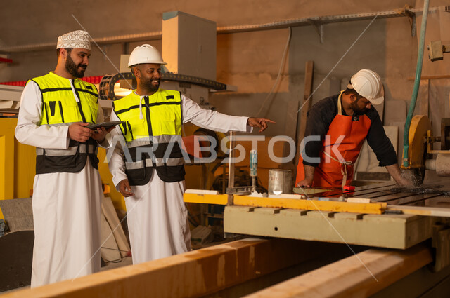 Monitoring and evaluating worker performance via tablet, factories and workshops in the Sultanate of Oman, marble manufacturing machines and equipment, quality of local national production, two Omani Gulf Arab engineers wearing dishdashas and work clothes working on tablets, integrating technology into factories