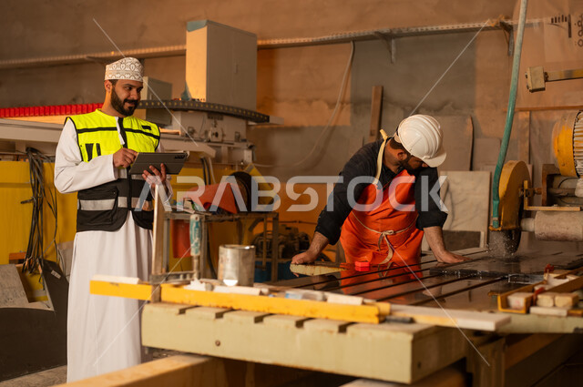 Monitoring and evaluating employee performance via tablet, factories and workshops in the Sultanate of Oman, marble manufacturing machines and equipment, quality of local national production, an Arab Gulf Omani engineer wearing a dishdasha and work clothes working on a tablet, integrating technology into factories.