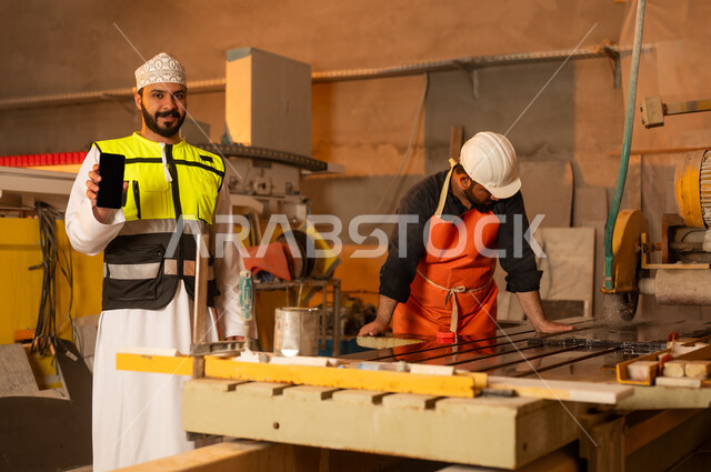 Worker performance check and evaluation, blank black screen on mobile phone, workshops in the Sultanate of Oman, tools and equipment made of marble, quality of local national production, home and office furniture industry, an Arab Gulf Omani engineer man wearing a dishdasha and work clothes holding a mobile phone in his hand