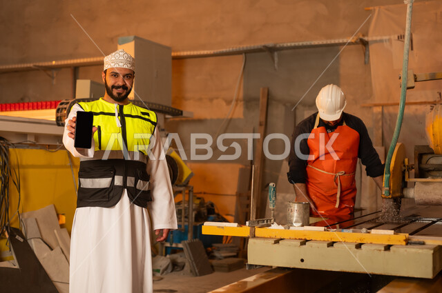 Worker performance check and evaluation, blank black screen on mobile phone, workshops in the Sultanate of Oman, tools and equipment made of marble, quality of local national production, home and office furniture industry, an Arab Gulf Omani engineer man wearing a dishdasha and work clothes holding a mobile phone in his hand