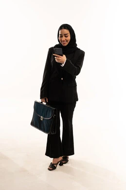 متابعة آخر المستجدات في العمل، دمج التقنية والتكنولوجيا في الحياة العملية، بورتريه لامرأة عربية سعودية خليجية محجبة ترتدي البدلة الرسمية السوداء تقف باستقامة و تحمل بيدها الموبايل و حقيبة اليد, ايماءات السعادة والسرور، خلفية بيضاء