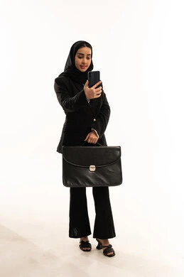 انجاز صفقات الاعمال عن طريق الموبايل, دمج التقنية والتكنولوجيا في الحياة العملية، بورتريه لامرأة عربية سعودية خليجية محجبة ترتدي البدلة الرسمية السوداء تقف باستقامة و تحمل بيدها الموبايل و حقيبة اليد, ايماءات السعادة والسرور، خلفية بيضاء