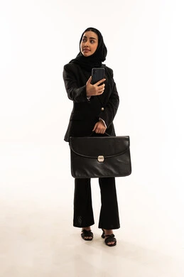 Going to work actively, the concept of business and project management, integrating technology into working life, a portrait of a veiled Saudi Arabian woman wearing a black formal suit, standing upright and holding a mobile phone and handbag, white background