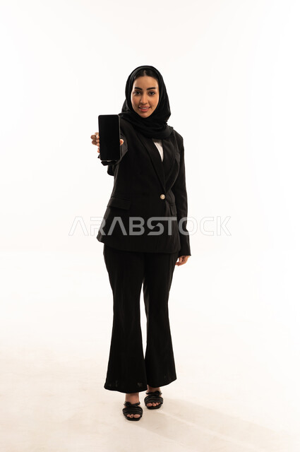 مشاركة الأخبار والأحاديث, التواصل مع العائلة والأصدقاء, استخدام الأجهزة التقنية الحديثة في الحياة اليومية,  بورتريه لامرأة عربية سعودية خليجية محجبة ترتدي البدلة الرسمية السوداء تقف باستقامة و تحمل بيدها الموبايل, خلفية بيضاء