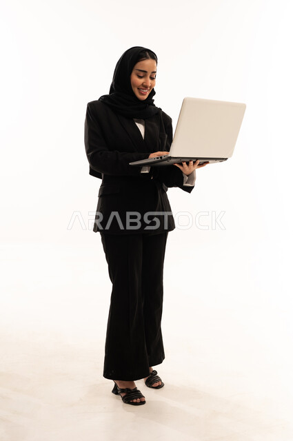 مشاهدة فيديوهات عبر الحاسب المحمول, استخدام الأجهزة التقنية الحديثة في الحياة اليومية, بورتريه لامرأة عربية سعودية خليجية محجبة مبتسمة ترتدي البدلة الرسمية السوداء تقف باستقامة و تحمل بيدها جهاز اللابتوب, خلفية بيضاء