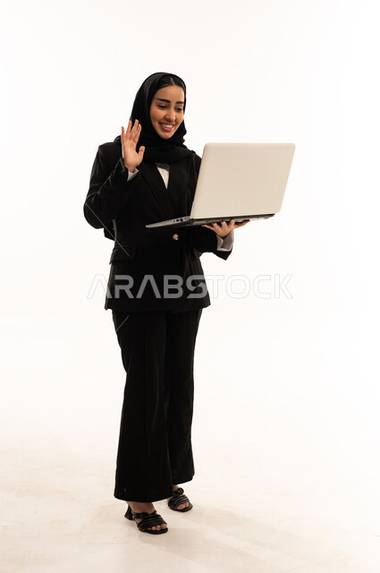 اجراء مكالمات الفيديو, استخدام الأجهزة التقنية الحديثة في الحياة اليومية, بورتريه لامرأة عربية سعودية خليجية محجبة مبتسمة ترتدي البدلة الرسمية السوداء تقف باستقامة و تحمل بيدها جهاز اللابتوب و ترفع يدها مرحبة, خلفية بيضاء